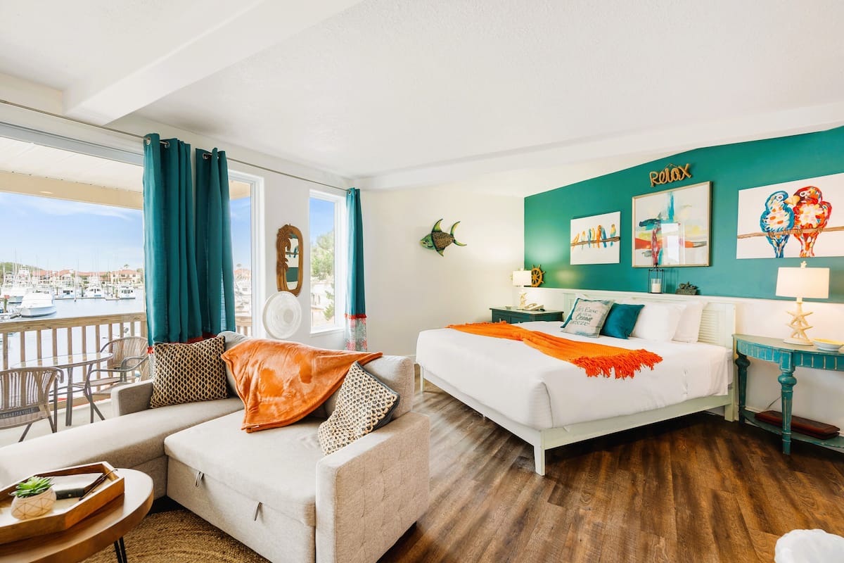 Una habitación de hotel moderna con balcón, una cama con una manta naranja, un sofá con cojines estampados y obras de arte en las paredes.