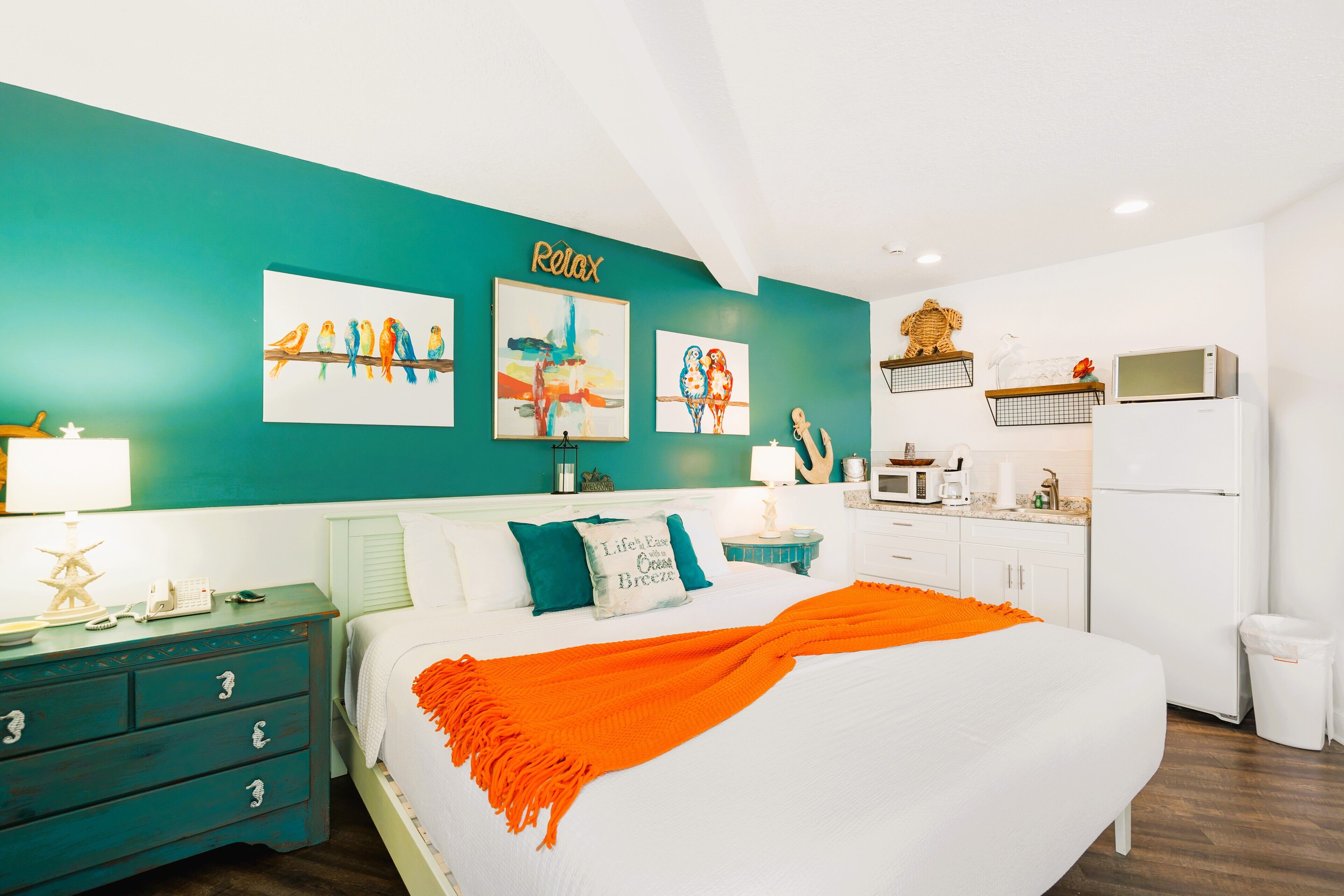 A bedroom with a bed, a nightstand, a dresser, a wall with artwork, a kitchen area with a microwave, and a refrigerator.