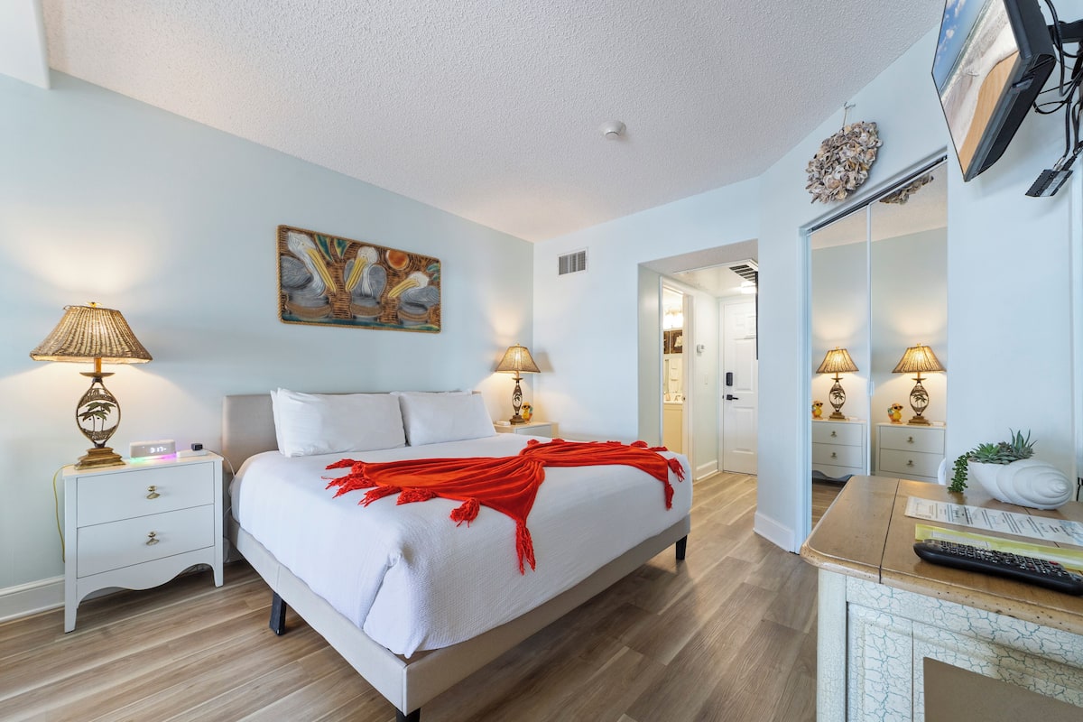 Un dormitorio con cama, lámparas de noche, mesita de noche, televisor y espejo.
