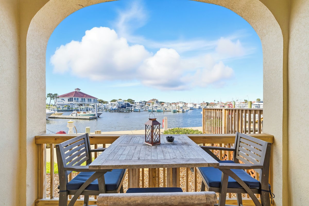 Una terraza de madera con una mesa y sillas, con vista a un puerto deportivo con barcos y un edificio con tejado rojo.