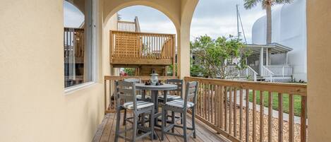 A wooden deck with a table and chairs, a wooden railing, and a view of a palm tree and building in the background.