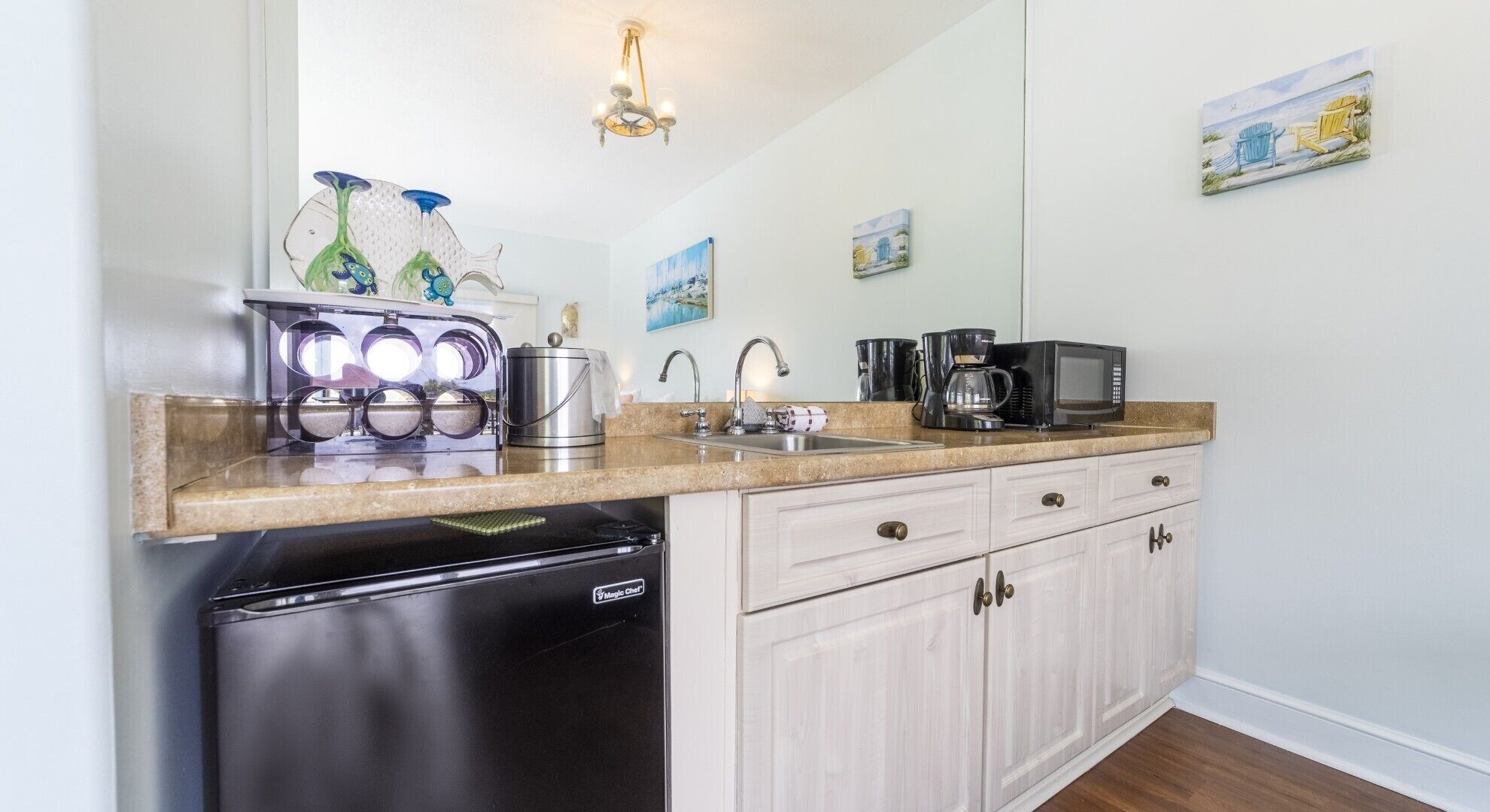 A compact kitchen with a sink, stove, and refrigerator. There are two wall-mounted light fixtures and a painting above the sink.