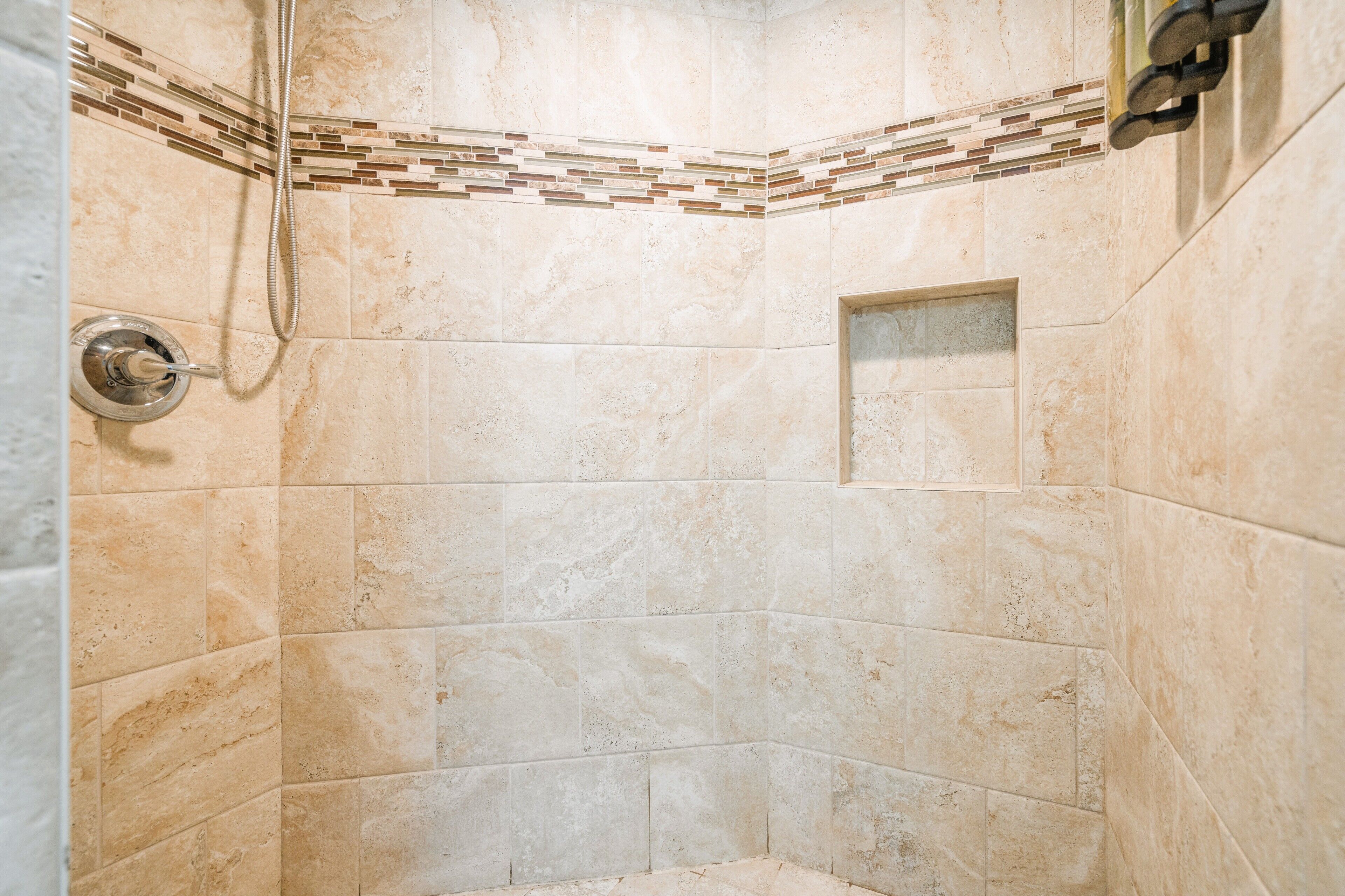 Un área de ducha con azulejos color beige, un cabezal de ducha y un diseño de azulejos en esquina.