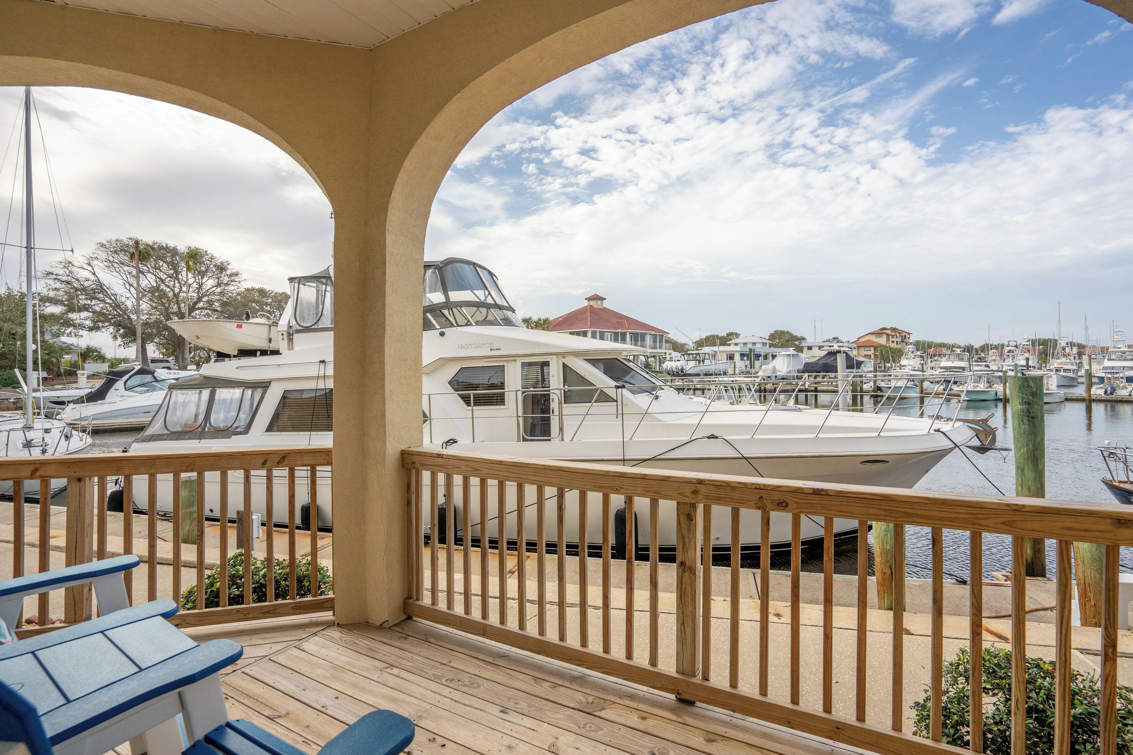 Une terrasse couverte, dotée d’une rambarde en bois, qui offre une vue sur une marina où sont amarrés plusieurs bateaux, avec un bâtiment visible au loin.
