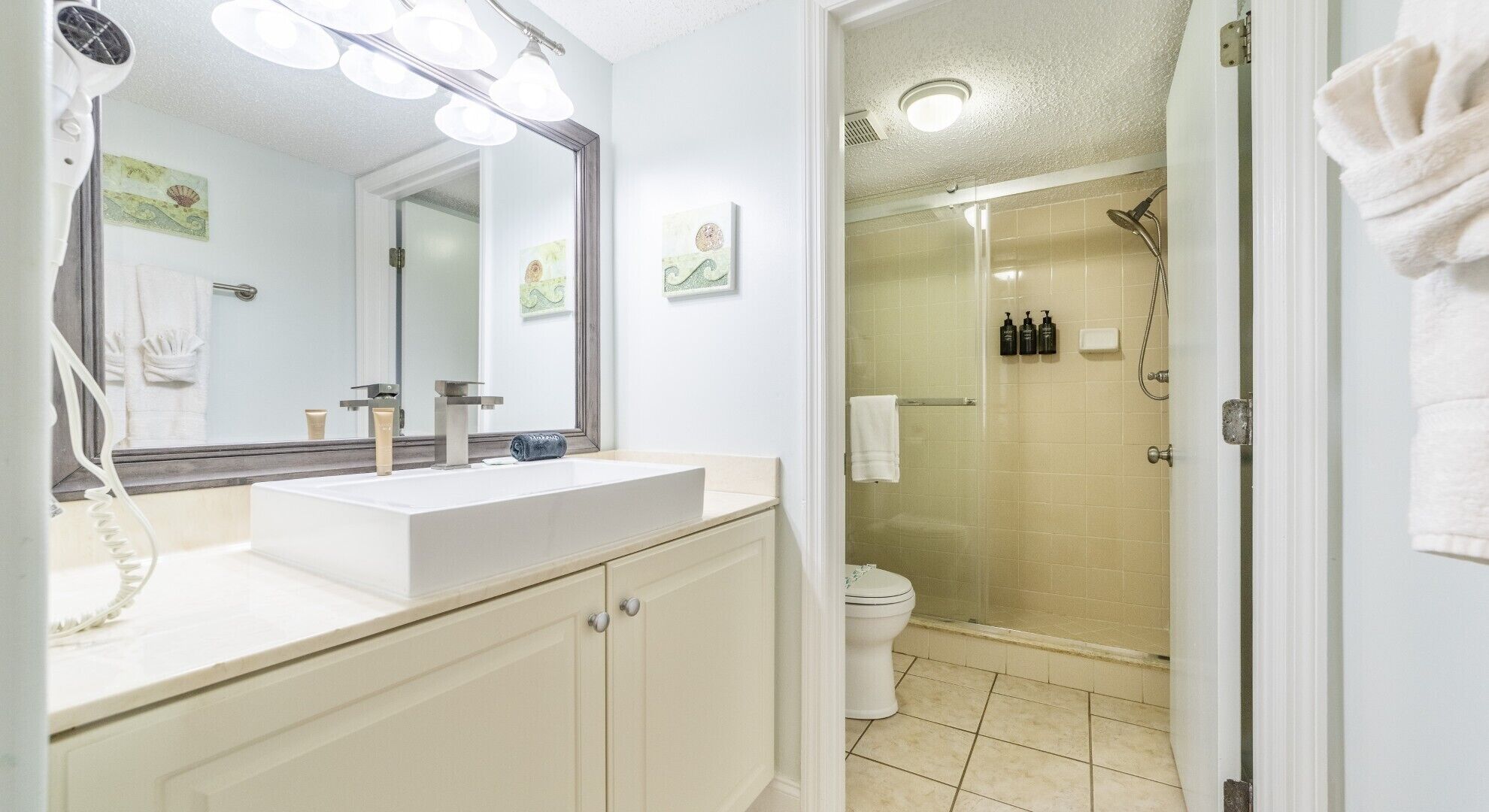 Une salle de bain avec un lavabo blanc, un miroir et une douche. On peut voir des serviettes accrochées au mur et des toilettes à l’arrière-plan.