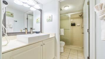 Une salle de bain avec un lavabo blanc, un miroir et une douche. On peut voir des serviettes accrochées au mur et des toilettes à l’arrière-plan.