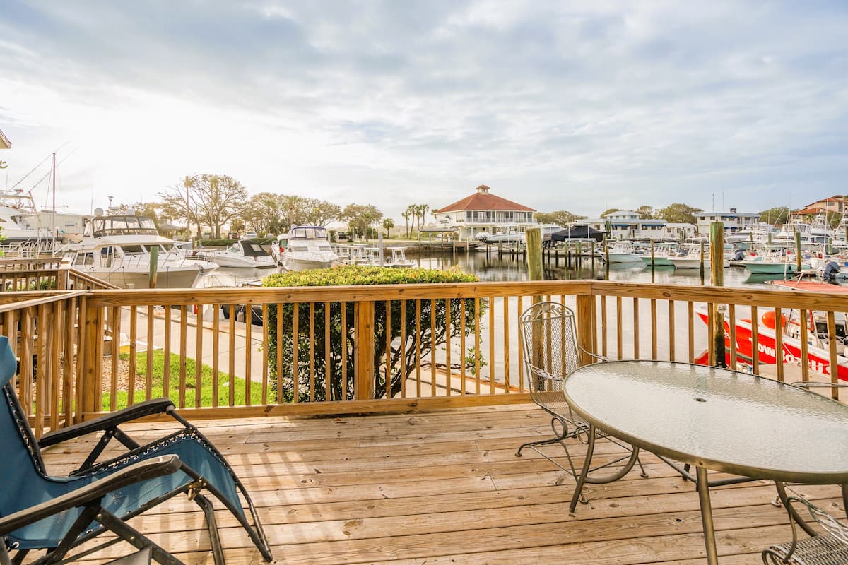Terraza de madera con mesa y sillas, con vista a un puerto deportivo con barcos y una casa al fondo.