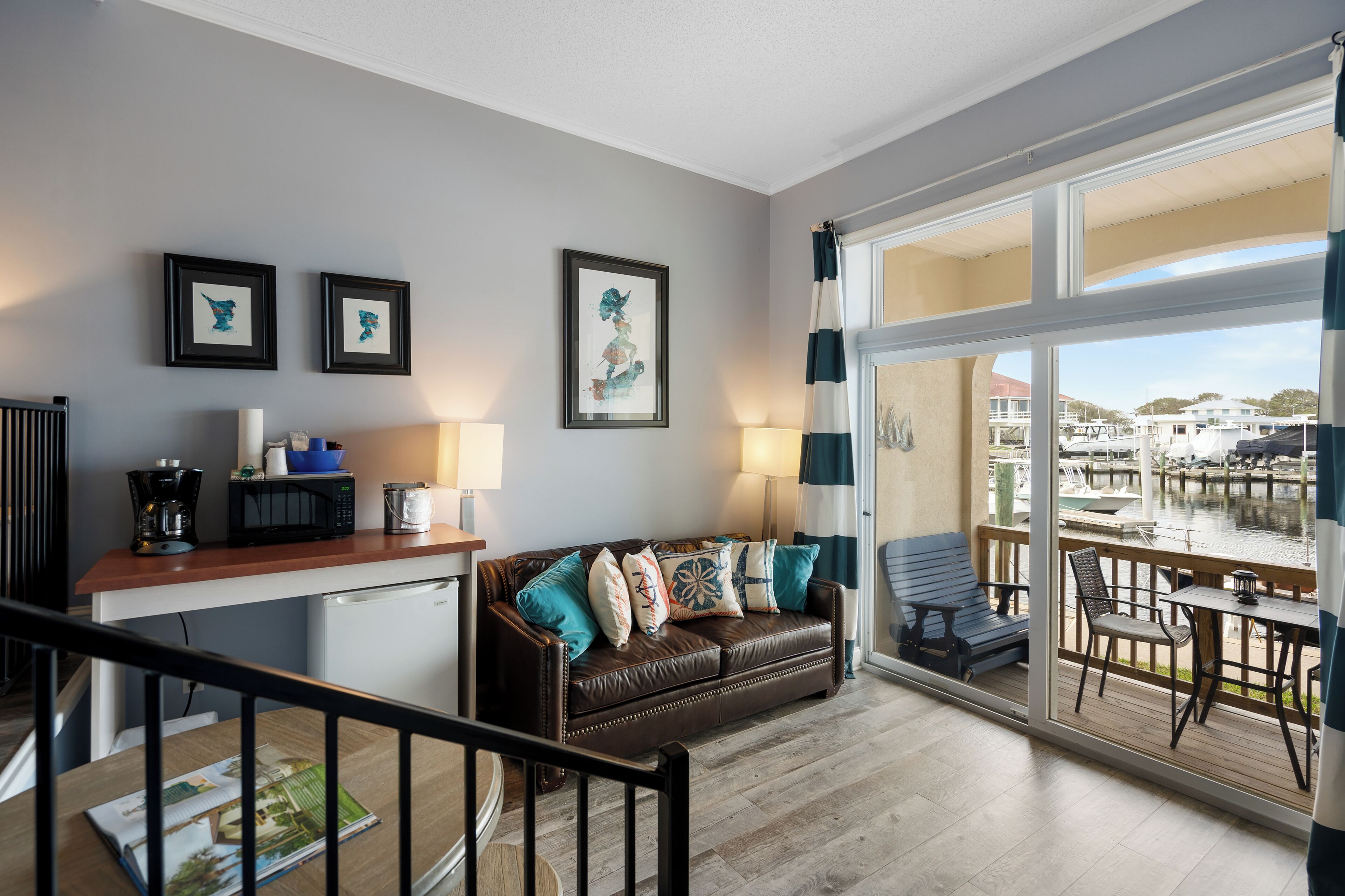 A modern living room with a brown leather sofa, a wooden coffee table, and a dining area with a table and chairs. There is a large window with a view of a marina.
