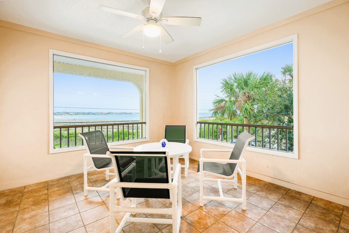 Una habitación con balcón, ventilador de techo, una mesa redonda con sillas y ventanas amplias que dan a vistas del mar y palmeras.