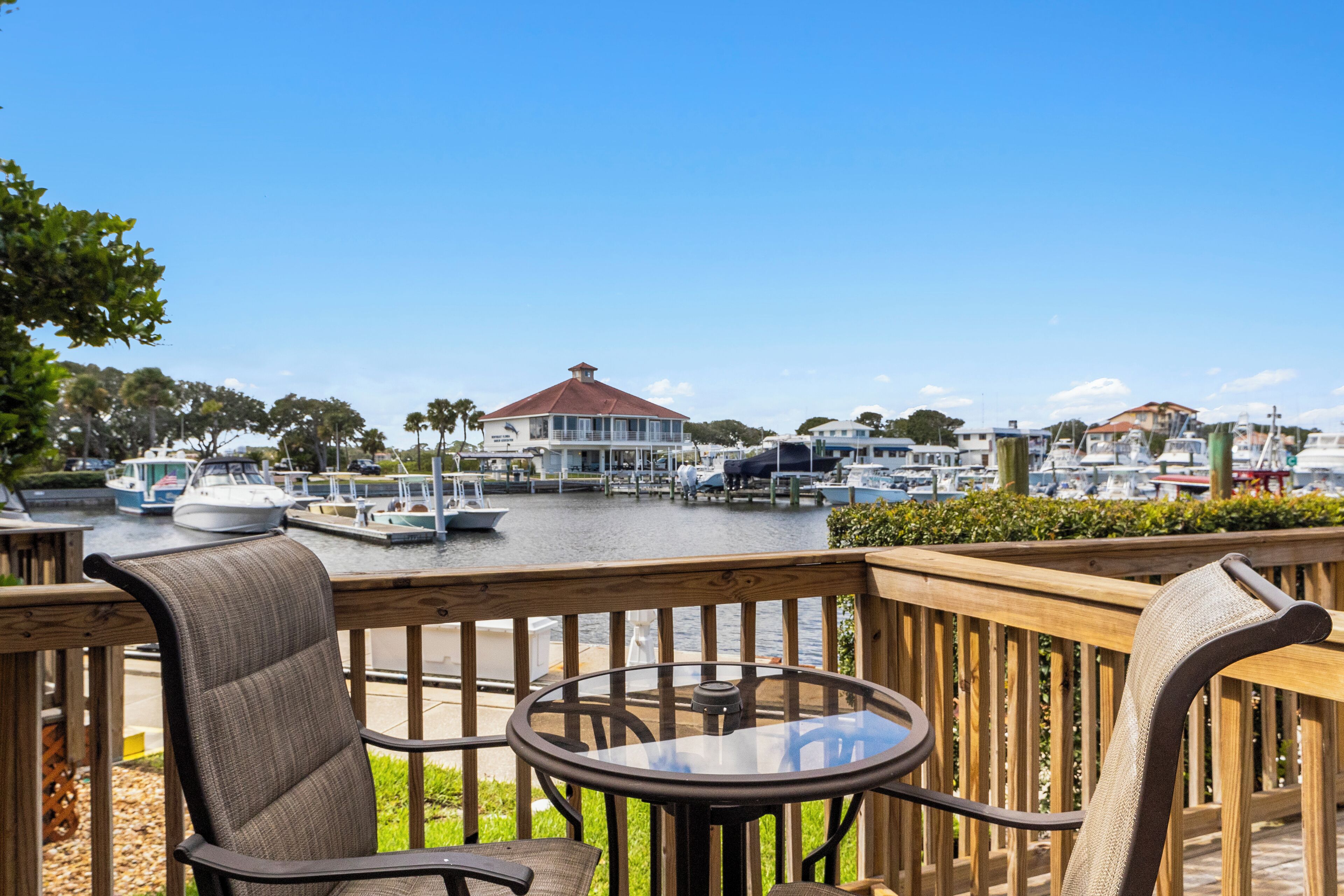 A wooden deck with a glass table and chairs overlooking a marina with boats and a building with a red roof.
