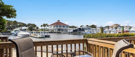 A wooden deck with a glass table and chairs overlooking a marina with boats and a building with a red roof.