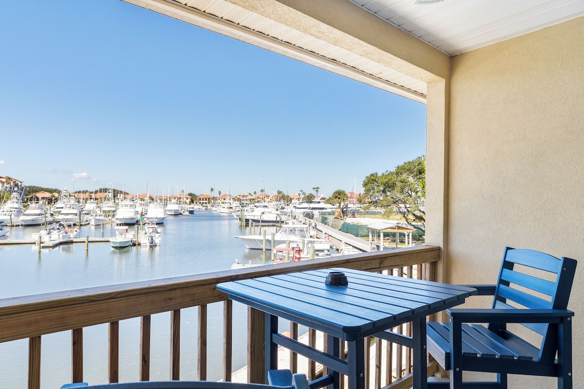Un balcón con vistas a un puerto deportivo lleno de barcos, barandilla de madera y una mesa con una silla.