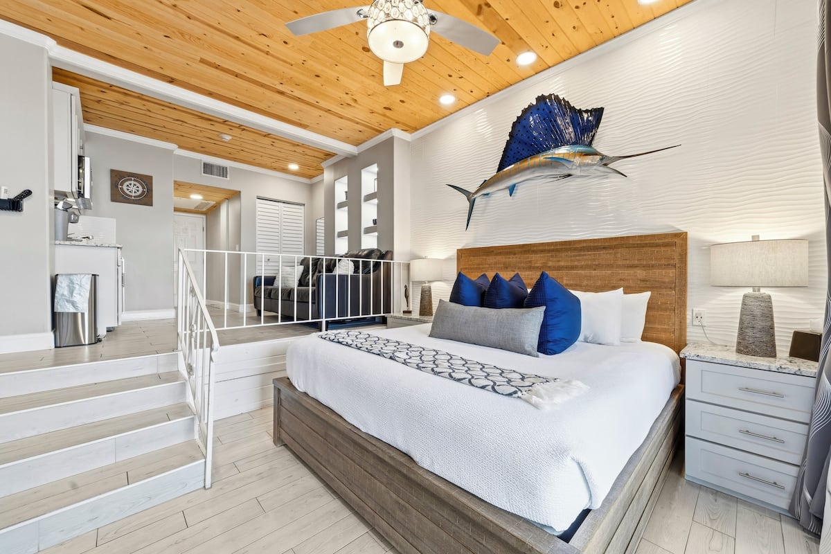 Un dormitorio moderno con techo de madera, una cama grande con ropa de cama estampada, una mesita de noche y una escultura decorativa de un pez en la pared.