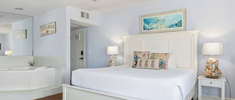 A bedroom with a large bed, a nightstand, a lamp, a painting, and a bathroom with a bathtub and a mirror.