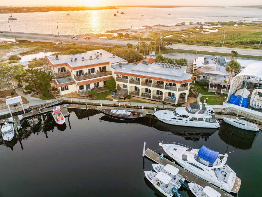 Uma marina com vários barcos atracados, um edifício grande com varandas e vista para a água e o pôr do sol.