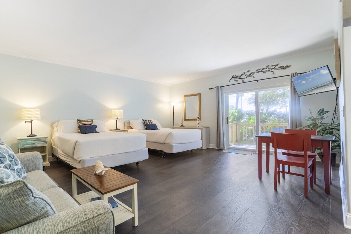 Habitación de hotel con dos camas, un sofá, una mesita, un escritorio con sillas y un balcón con vistas.