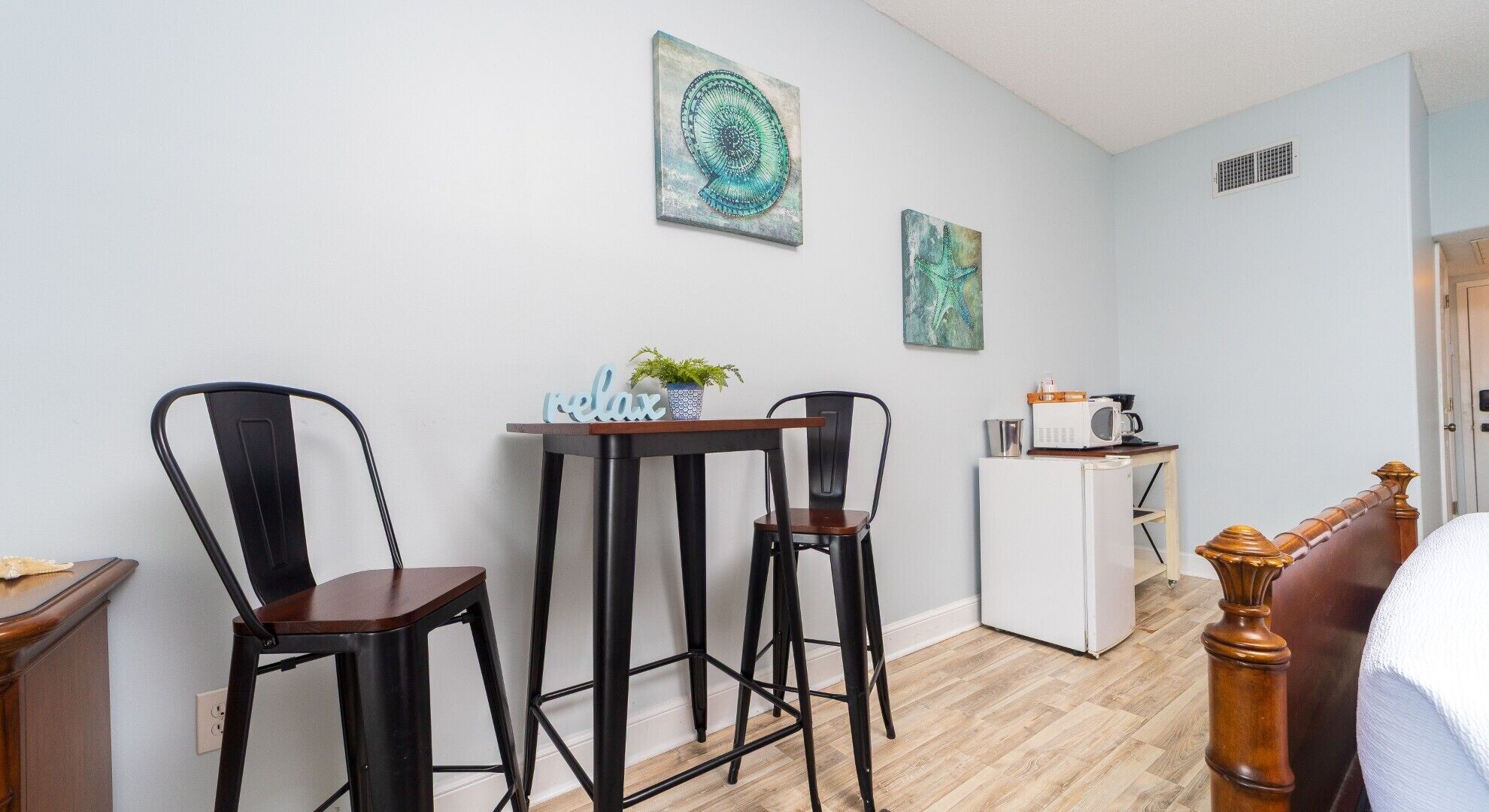 A small, modern room with a wooden floor, a white refrigerator, a microwave, and a bed with a wooden headboard. There are two black metal chairs and a small table with a plant and a sign that reads "relax".