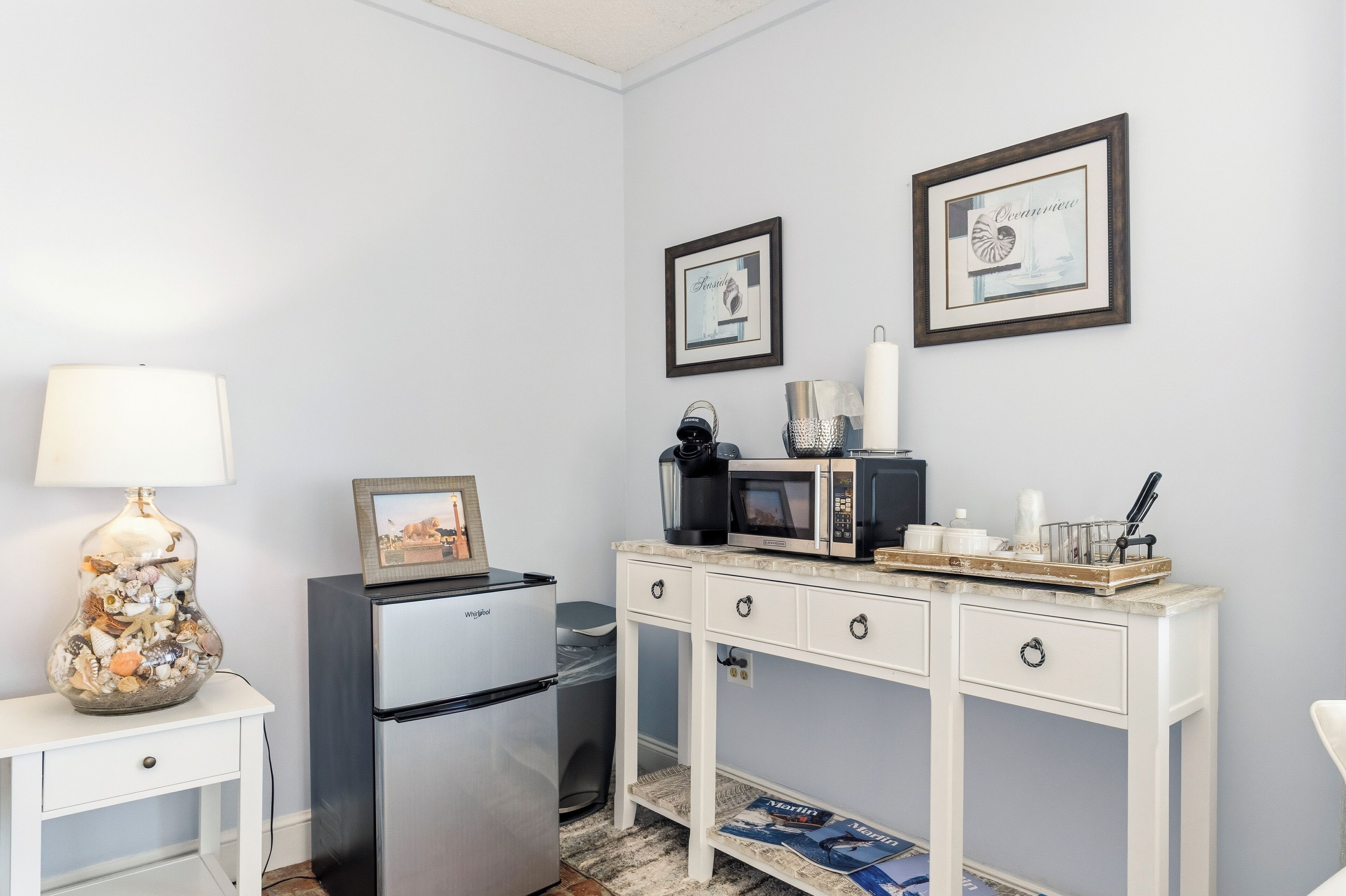 A compact kitchenette with a refrigerator, microwave, and a small dining table with a lamp and decorative items.