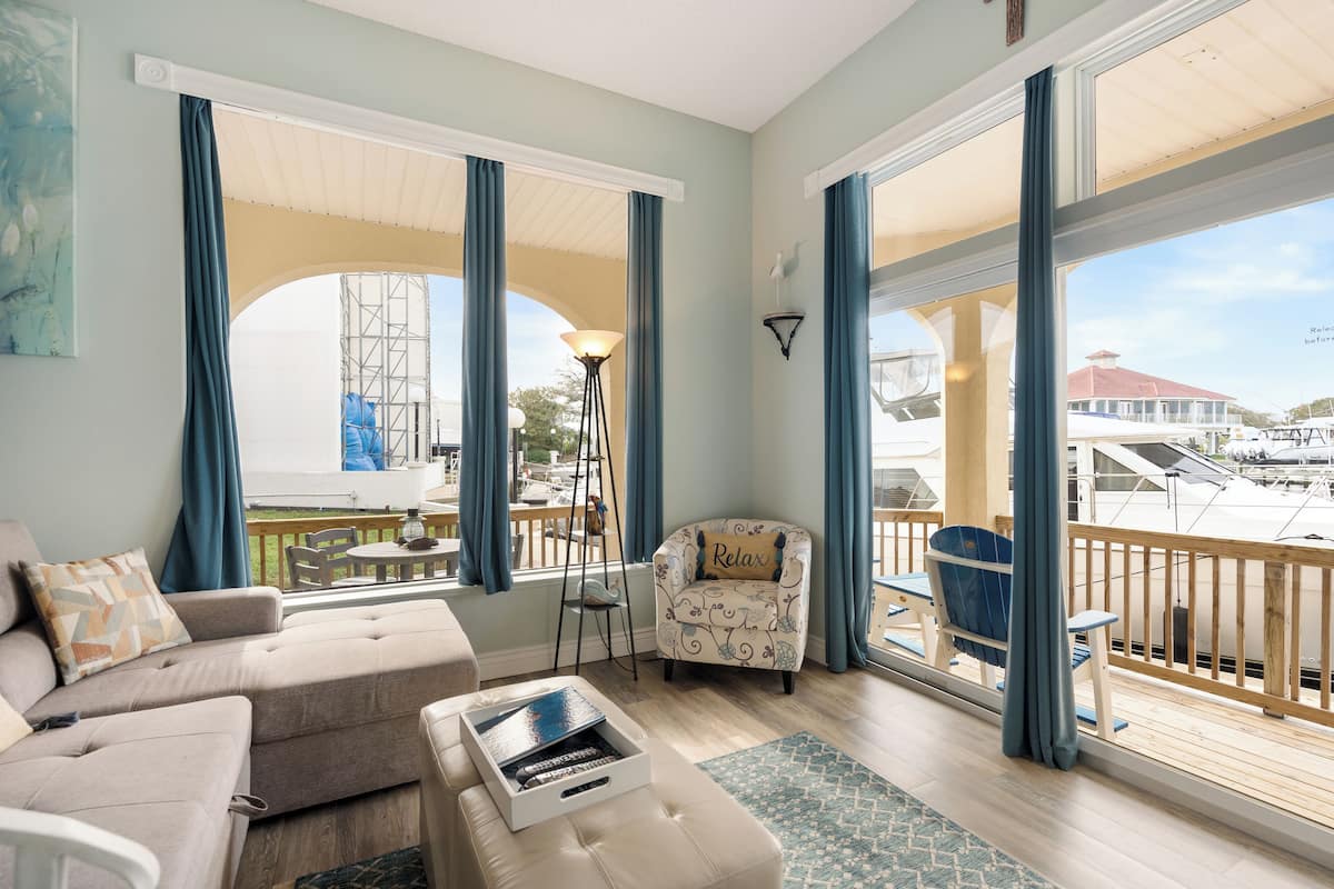 Amplia sala de estar con sofá, sillón y una mesita con bandeja. Grandes ventanales ofrecen vistas a un puerto deportivo con barcos y un edificio.