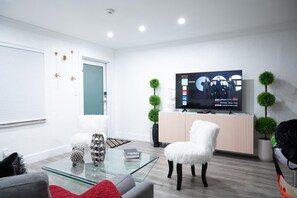 A modern living room with a glass coffee table, a white chair, a television on a stand, and decorative plants.