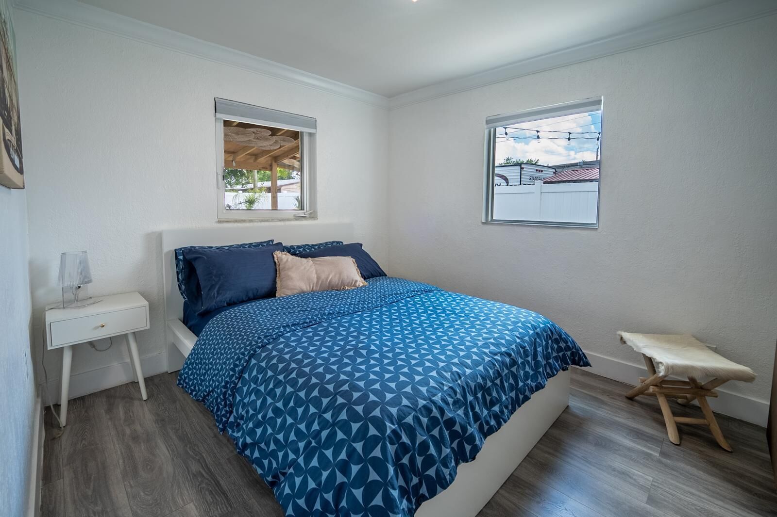 Een slaapkamer met een bed, nachtkastje, raam en een klein krukje.
