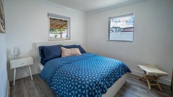 Een slaapkamer met een bed, nachtkastje, raam en een klein krukje.