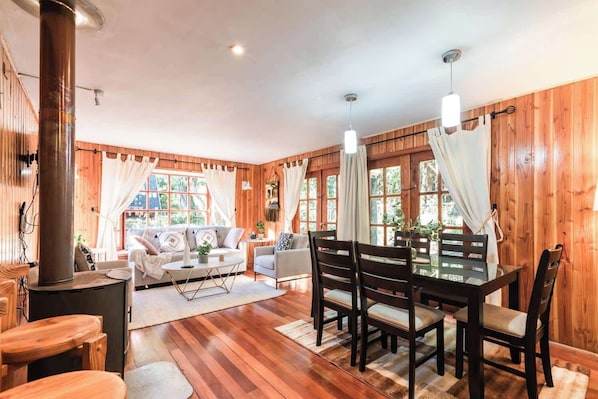 A cozy room with a wooden interior, a dining table with chairs, a sofa, and a large window with curtains.