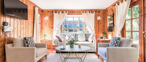 A cozy room with wooden walls, a flat-screen TV, two sofas, a coffee table, and a large window with curtains.