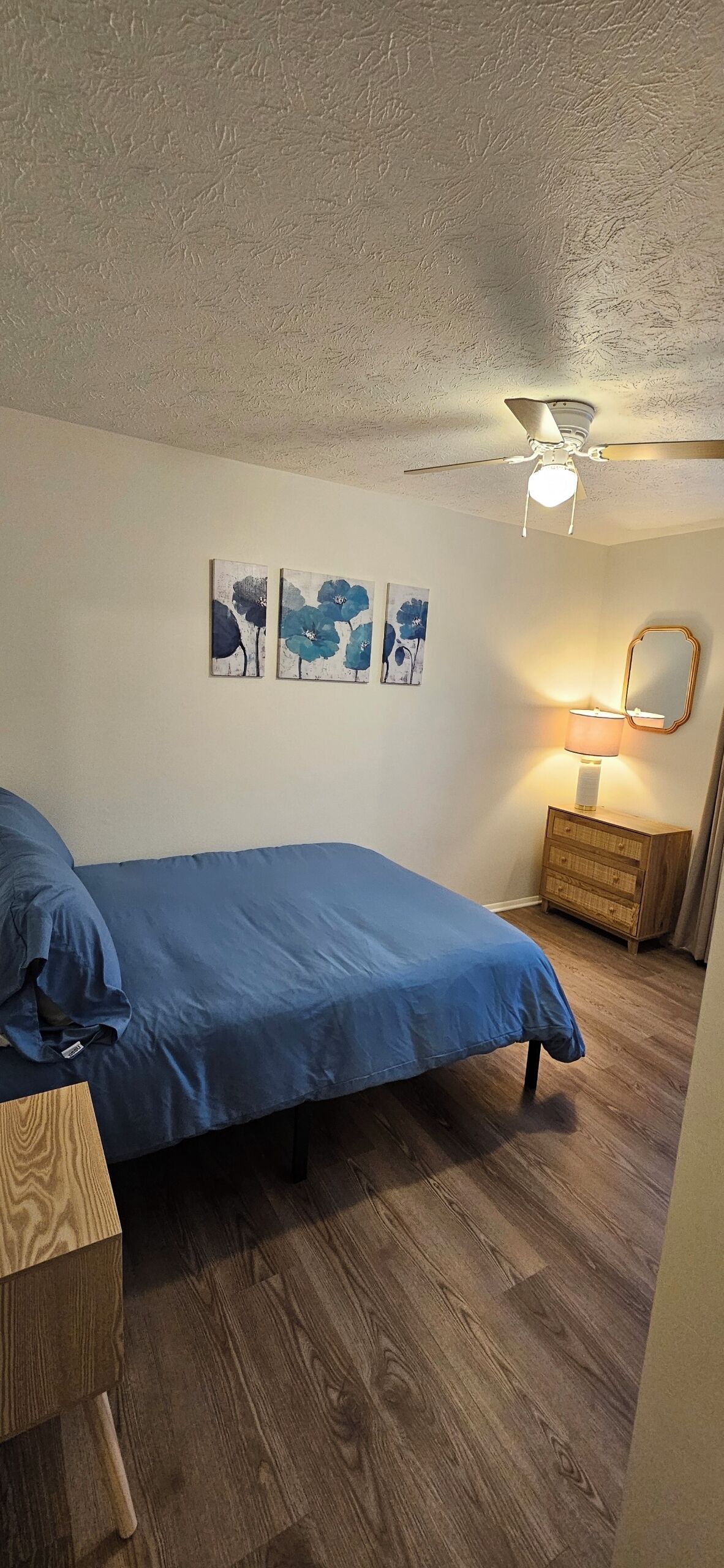 A bedroom with a bed, wooden floor, ceiling fan, wall-mounted lamp, and artwork.