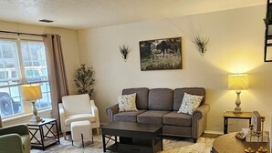 Un salón con suelo de madera, un sofá gris, un sillón blanco, una mesa de centro y una lámpara.