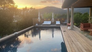 Un espace piscine agrémenté de chaises longues et de parasols, entouré d’une terrasse en bois et de verdure, offrant une vue sur un paysage montagneux.