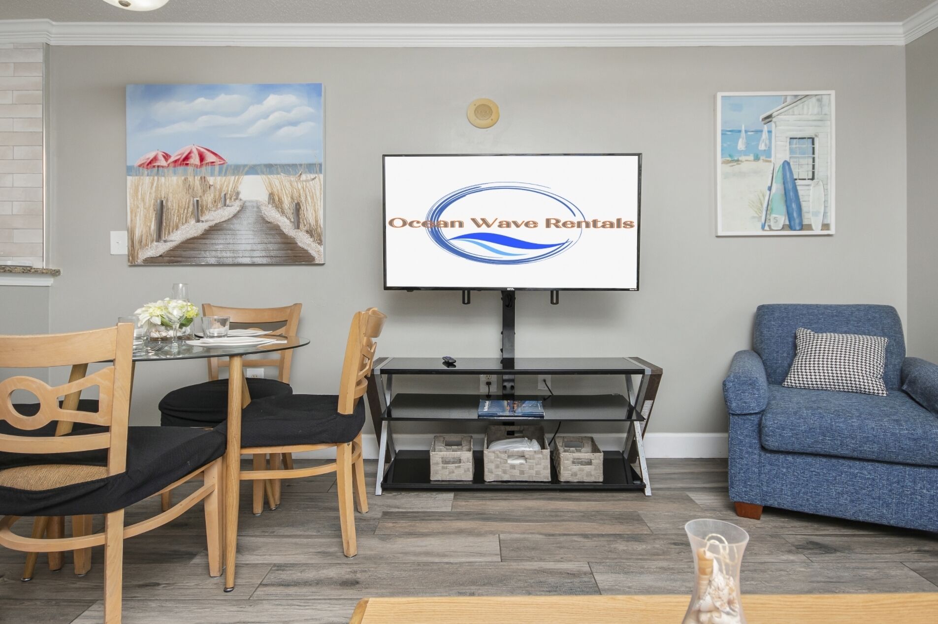 A living room with a TV screen displaying 'Ocean Wave Rentals,' a dining area with chairs and a table, and a blue sofa.