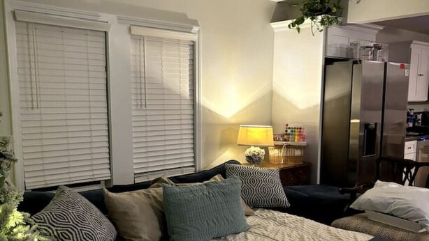 A bedroom with a bed, pillows, a nightstand, a lamp, a window with blinds, and a kitchen area with a refrigerator.