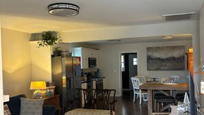 A spacious living room with a dark wood floor, a blue sofa, a dining area with a wooden table, and a kitchen in the background.
