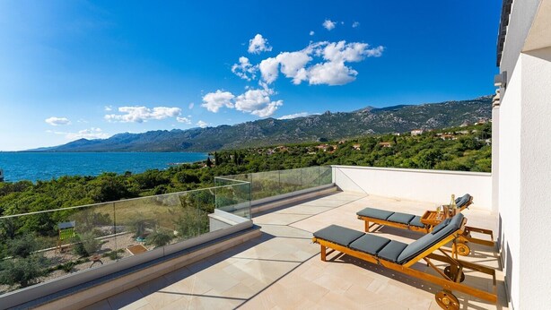 Un balcon doté de deux chaises longues et d’une balustrade en verre, offrant une vue panoramique sur un lac et des montagnes.
