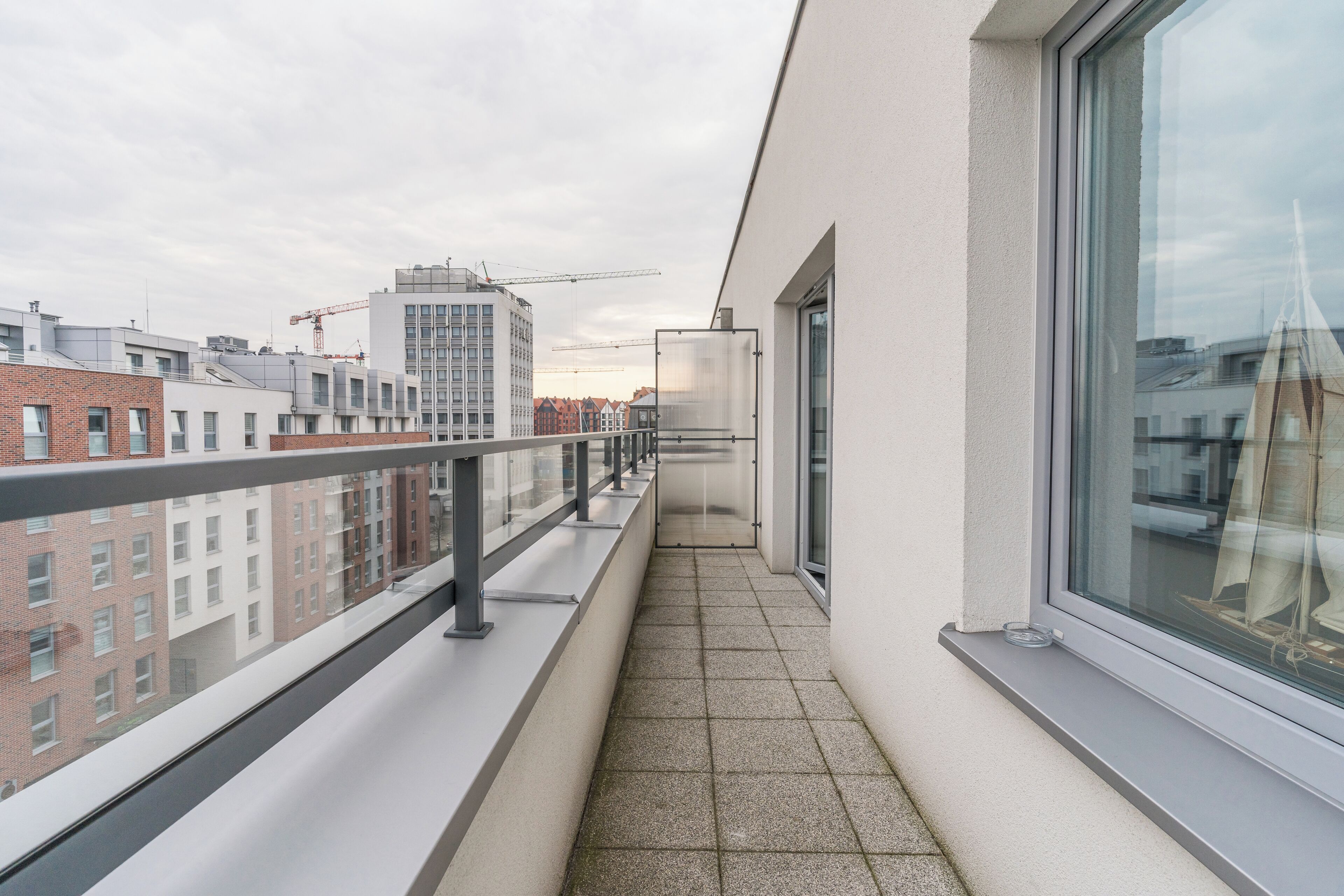 Ein schmaler Balkon mit Glastrennwand, Fliesenboden und Blick auf umliegende Gebäude sowie einen Kran in der Ferne.
