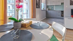 A modern kitchen with white cabinets, a dining area with a table set for two, and a balcony with a plant and curtains. - Langgarten - Little Heaven Apartamenty (Gdansk)
