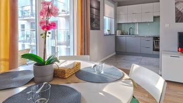 A modern kitchen with white cabinets, a dining area with a table set for two, and a balcony with a plant and curtains.