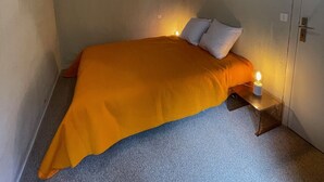 Et lille enkeltværelse med en simpel lysarmatur over sengen og en dør på højre side.