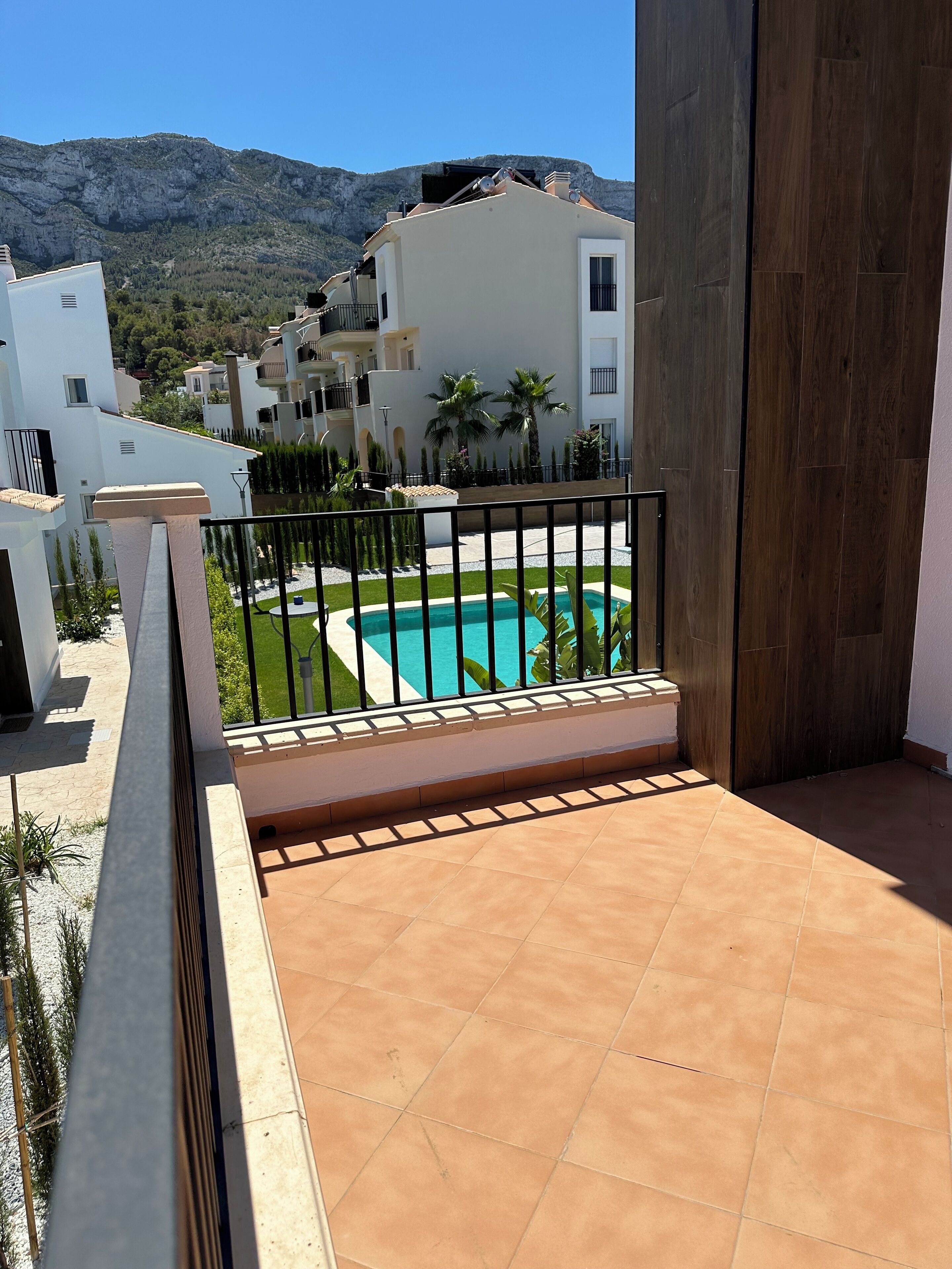 A balcony with a view of a swimming pool, surrounded by white buildings and a mountain in the background.
