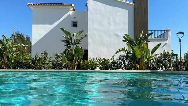 A swimming pool with clear water, surrounded by lush greenery and a white building with a tiled roof.