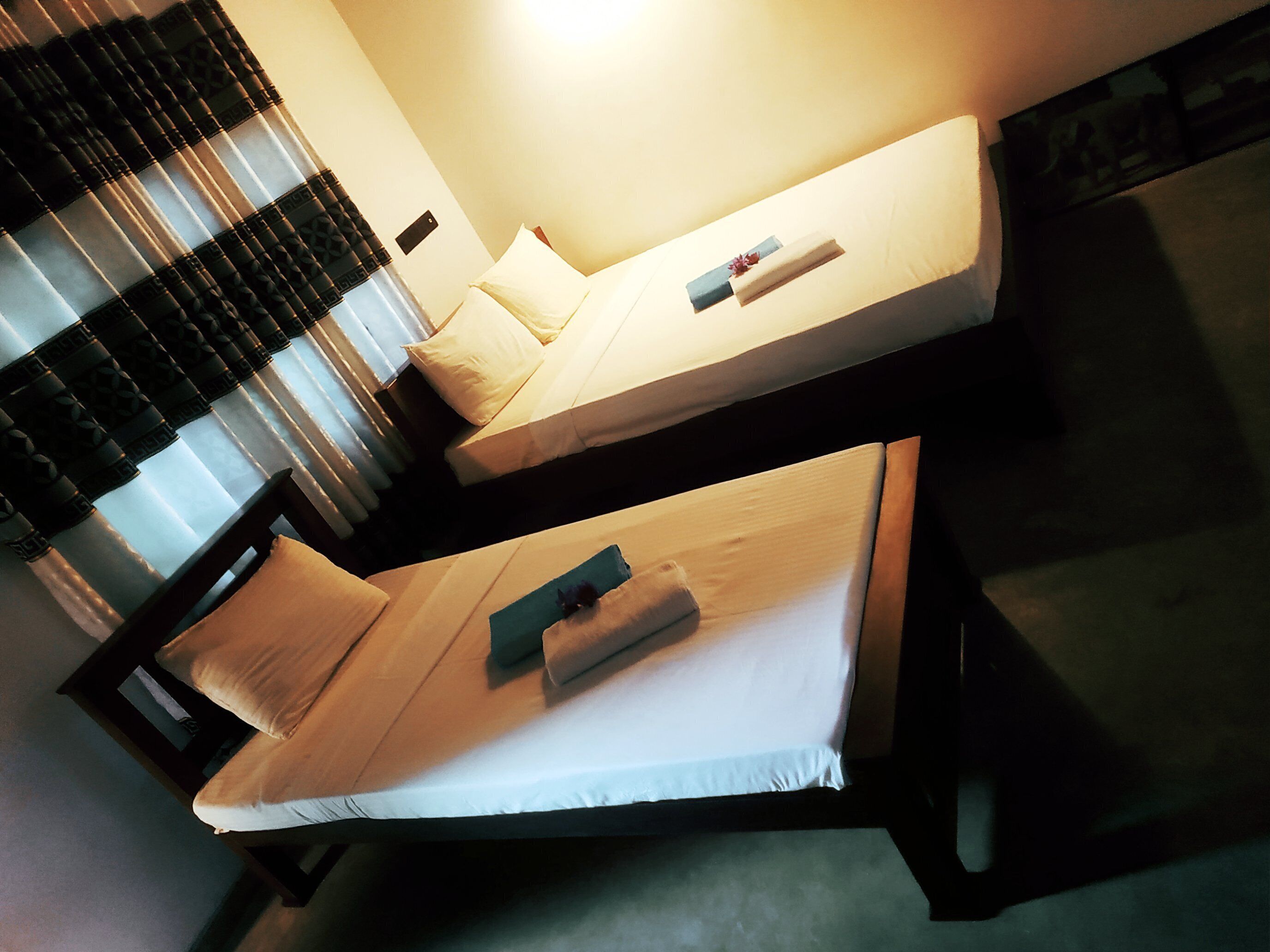 Two single beds with pillows and a blanket, a window with blinds, and a wall-mounted light fixture.