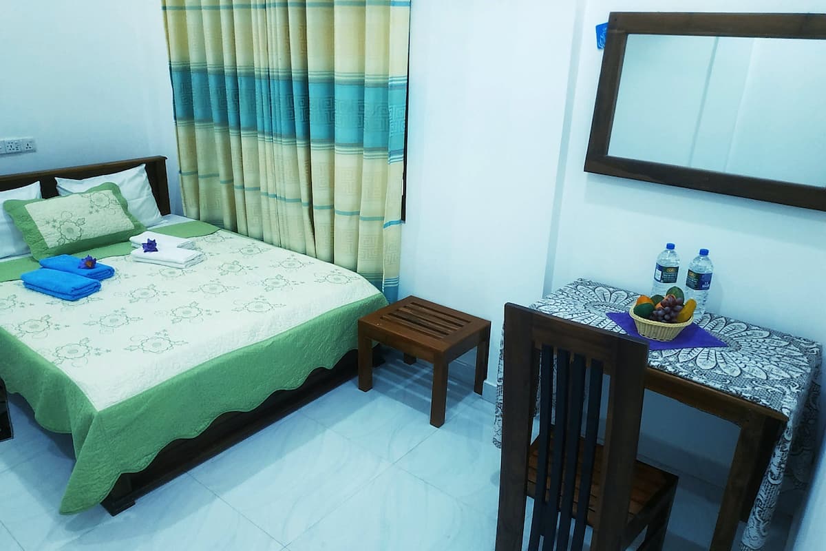 Una camera da letto ordinata con un letto, un piccolo tavolino di legno con una tovaglia decorata e una sedia. Sul tavolo ci sono due bottiglie d'acqua.