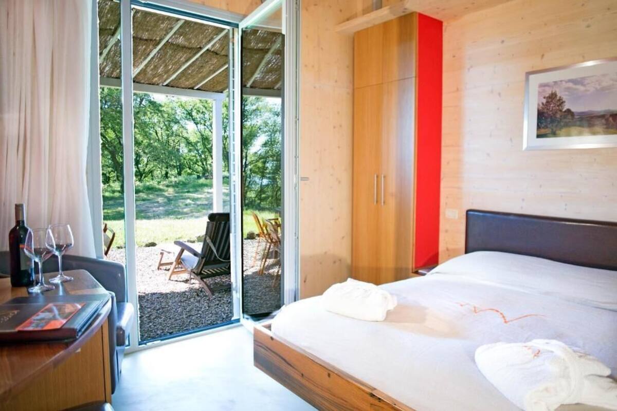 A bedroom with a bed, a desk with a bottle and glasses, a red cabinet, and a view of the outdoors.