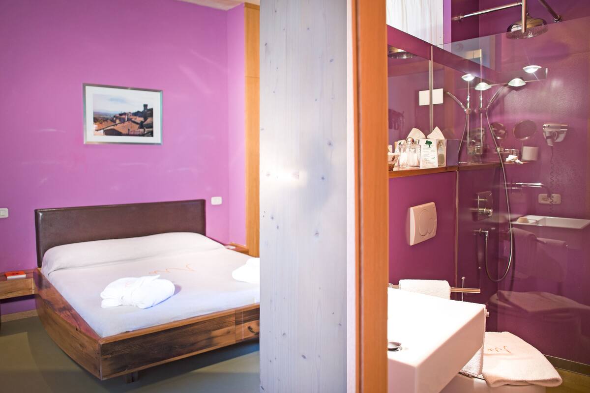 A room with a wooden bed, a bathroom with a shower, and a purple wall.
