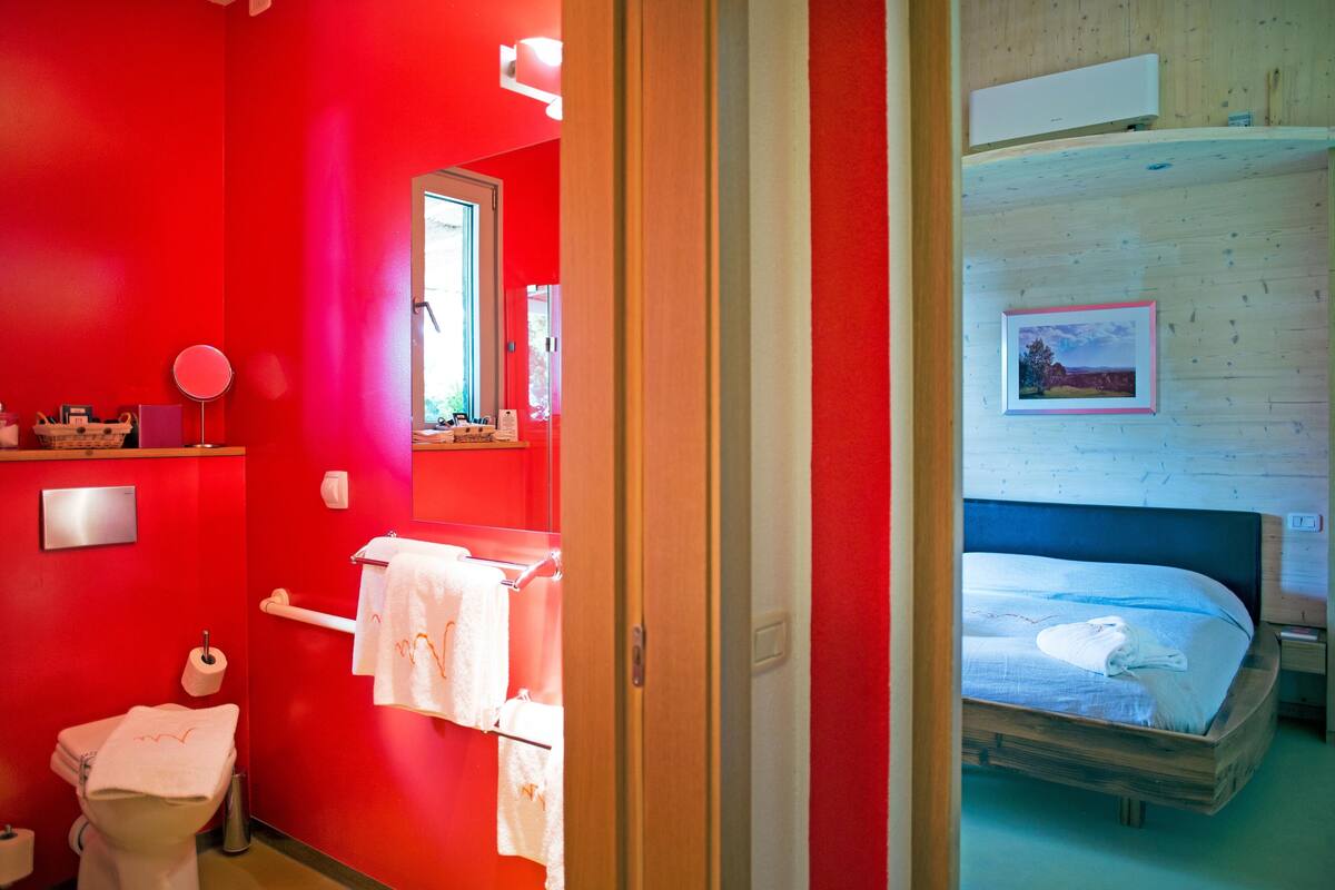 A room with a red wall, a wooden bed frame, a white toilet, and a small window.