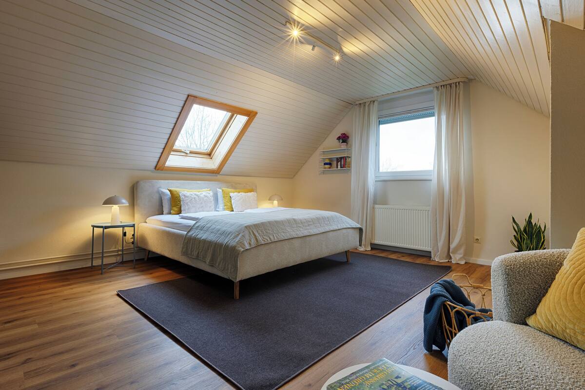 Ein Schlafzimmer mit Bett, Nachttisch mit Lampe, einem Fenster mit Vorhängen, einer Pflanze und Blick ins Freie.