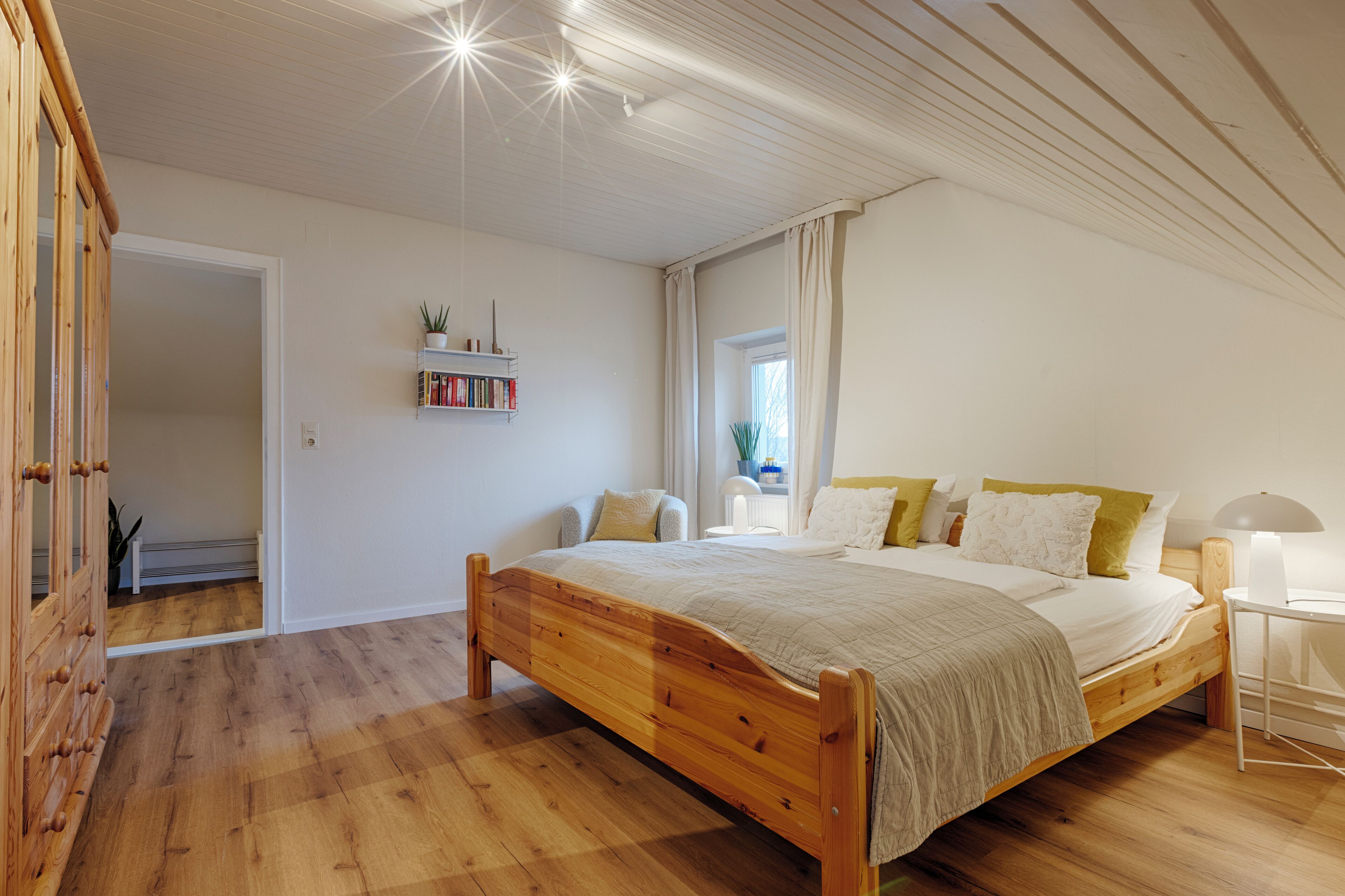 Une chambre à coucher avec un lit en bois, une table de chevet, une lampe, une étagère remplie de livres et une fenêtre avec des rideaux.