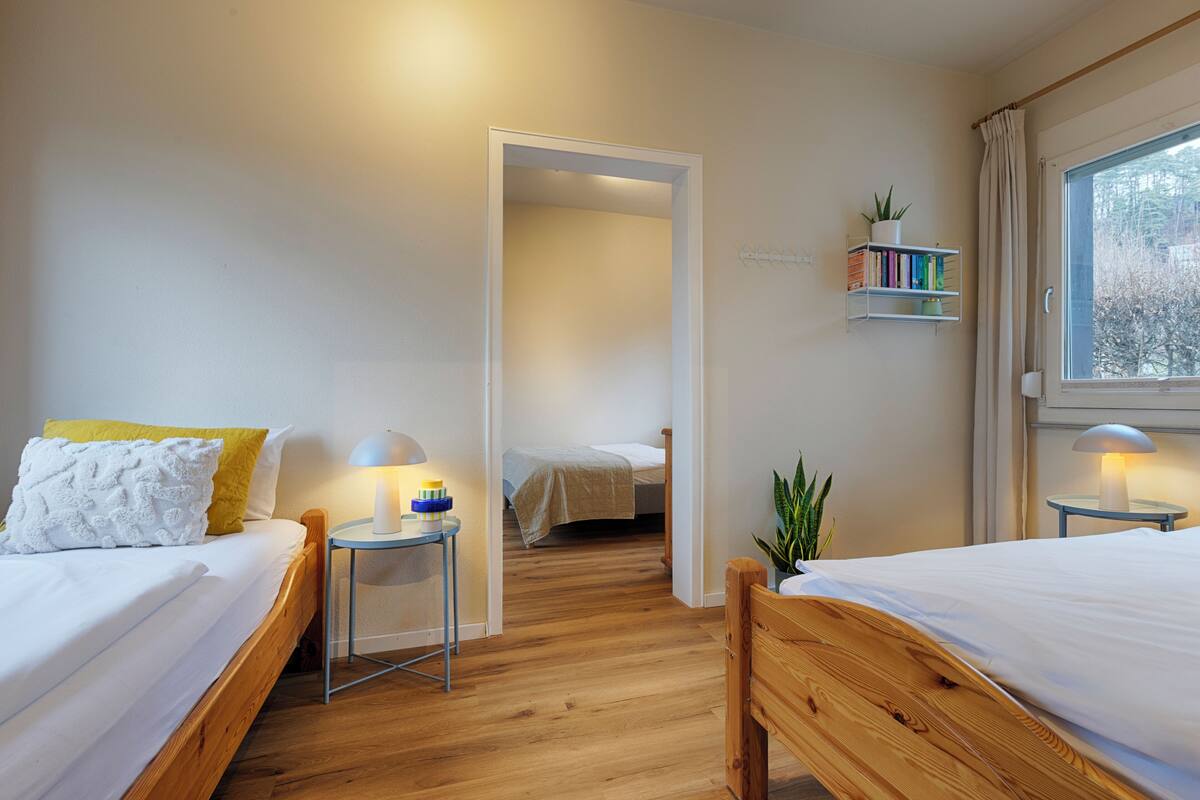Ein Schlafzimmer mit zwei Betten, einem Nachttisch mit Lampe, einem Bücherregal und einem Fenster mit Vorhängen.