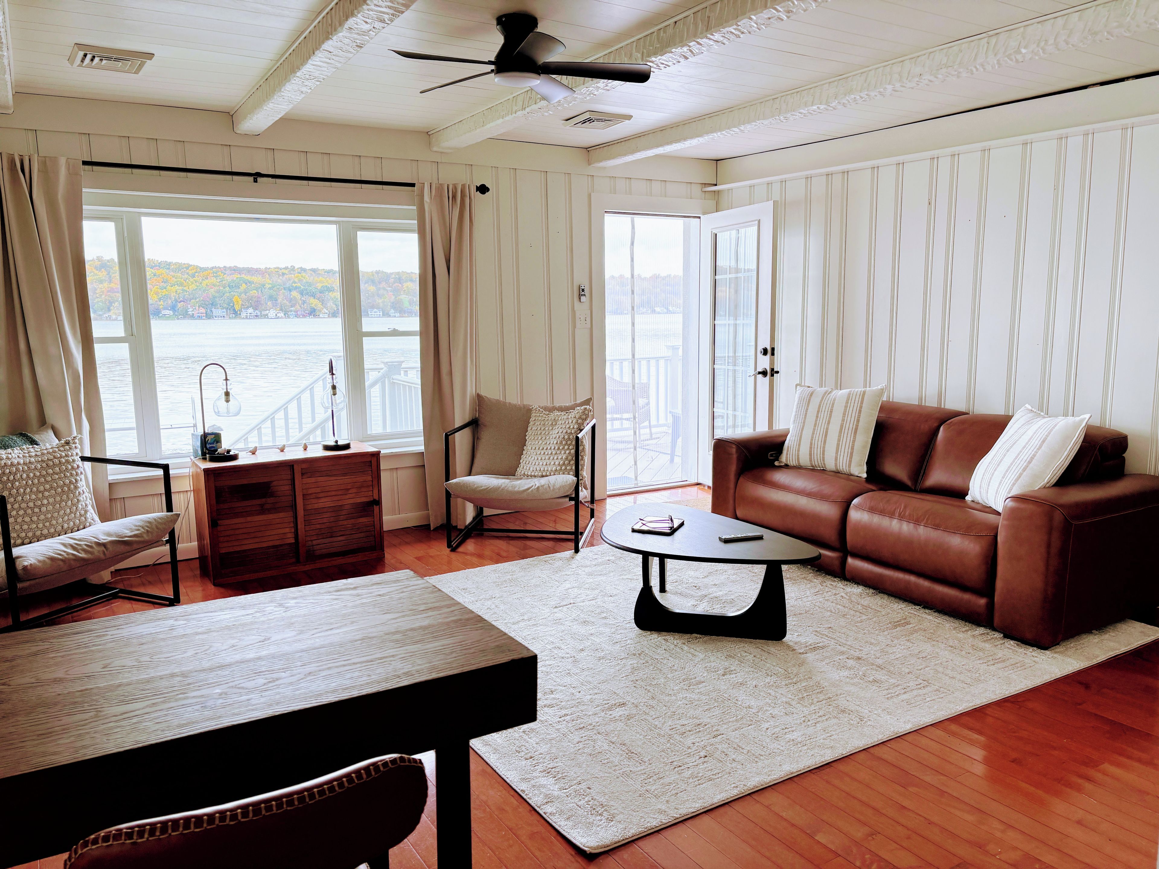 A living room with a brown leather sofa, a wooden coffee table, and a chair with a cushion. There is a window with curtains, a ceiling fan, and a view of a body of water.